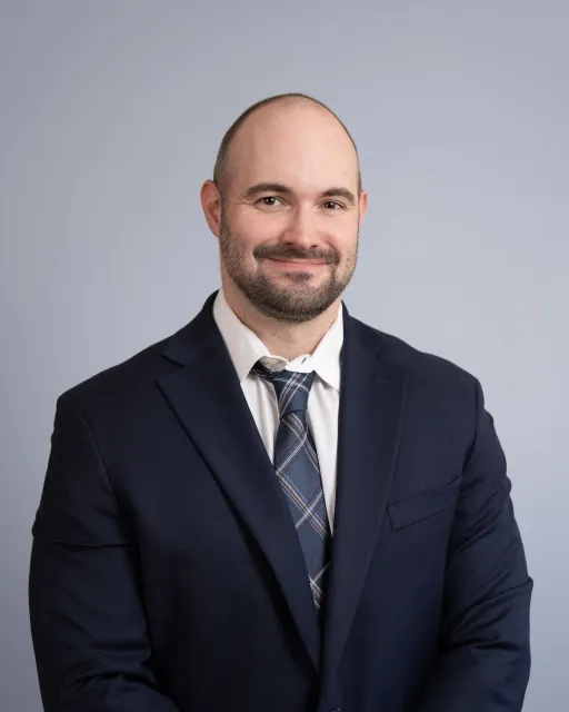 Professional portrait of a man with a beard and bald head, wearing a dark suit, white shirt, and plaid tie, posing against a plain gray background.