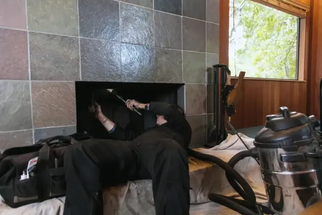 A person cleaning a fireplace, with a vacuum, tools, and a window in the background.