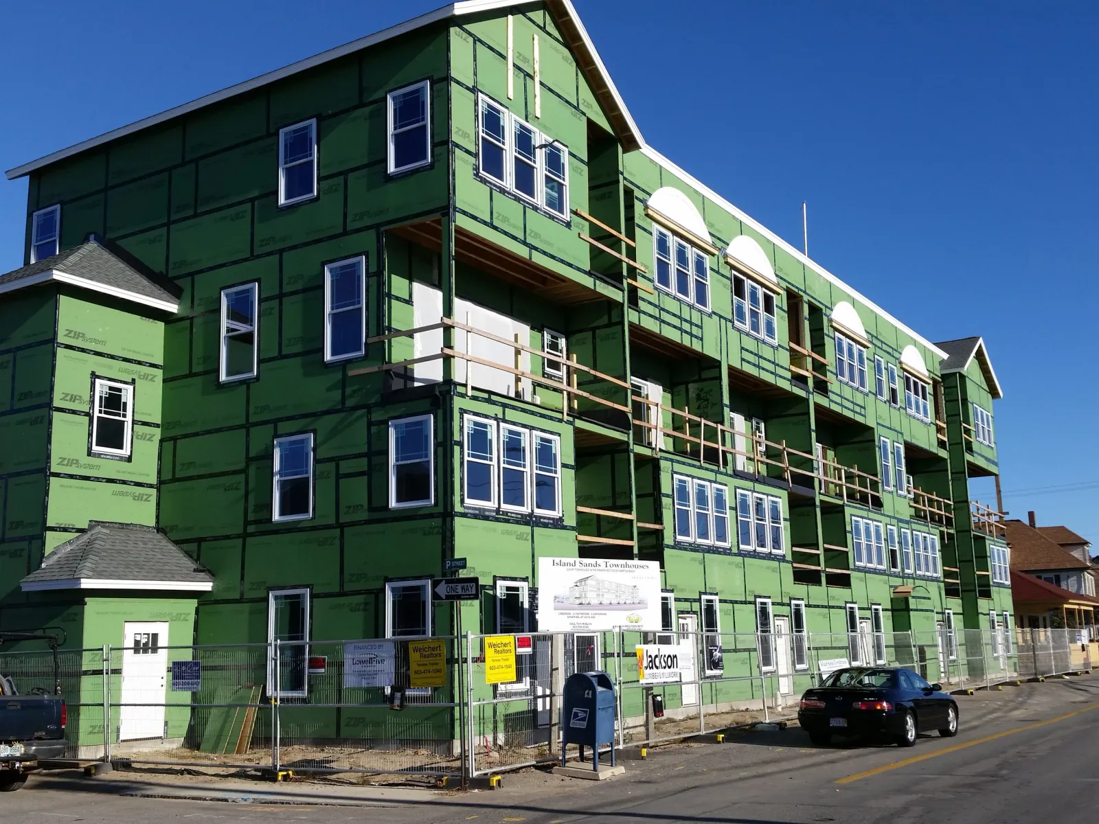 Green building under construction with multiple windows and balconies, surrounded by a temporary fence, parked cars, and street signs, against a clear blue sky.