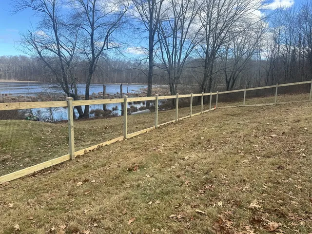 Single-rail pressure-treated wood fence with wire mesh running along a hillside beside a pond
