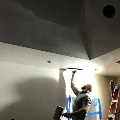 A person is plastering a large, angled ceiling in a dimly lit room. They are holding a plastering trowel and wearing a gray shirt.