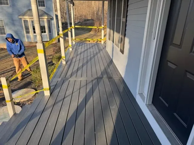 Gray deck under construction beside a house, with yellow caution tape and a worker in a blue hoodie nearby