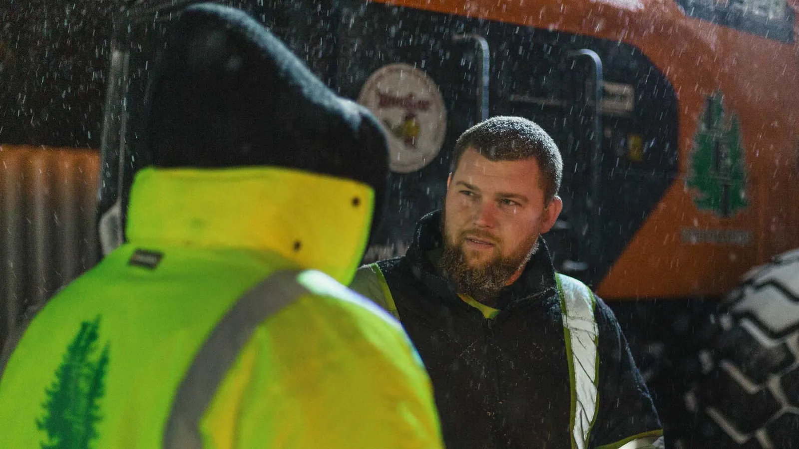 Two workers in rainwear talk outside at night with snow falling around them.