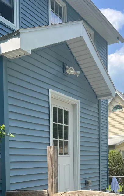 Blue-sided house with white door and awning; bright sunlight.