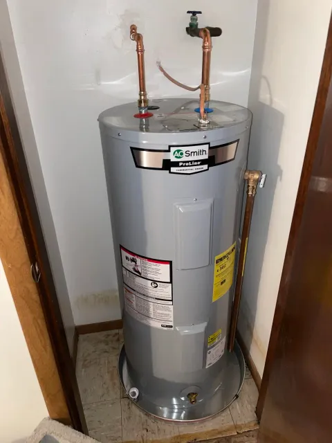 A gray electric water heater with copper pipes and a blue and red valve connected to the plumbing, positioned in a corner against a white wall.