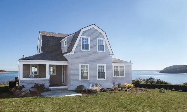 A two-story house with gray siding, white trim, and a large lawn overlooking the water and distant land.