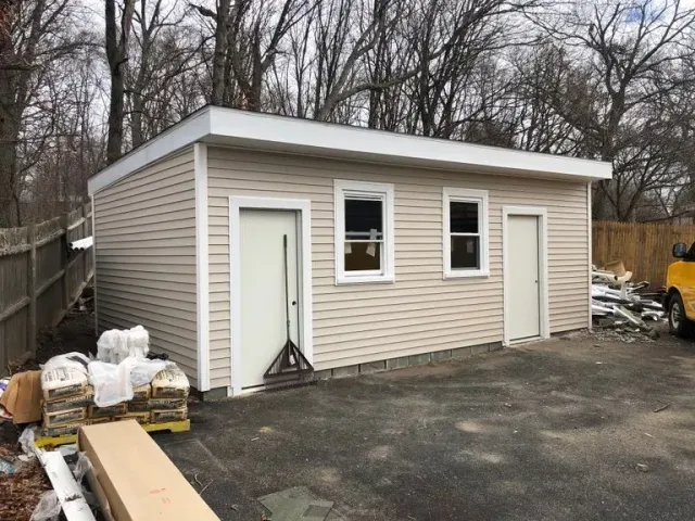 Small tan garage with two white doors, set on a paved lot beside a fence and leafless trees.