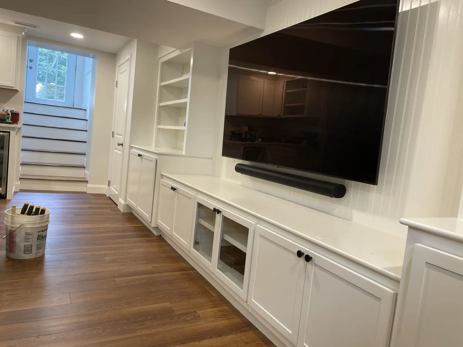 Living room with white built-in cabinets, a large flat-screen TV, and a wooden floor. A window and staircase are visible in the background. A bucket with tools or markers is on the floor.