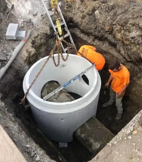 Workers installing a large concrete sewer well in an excavation site with construction tools and equipment.