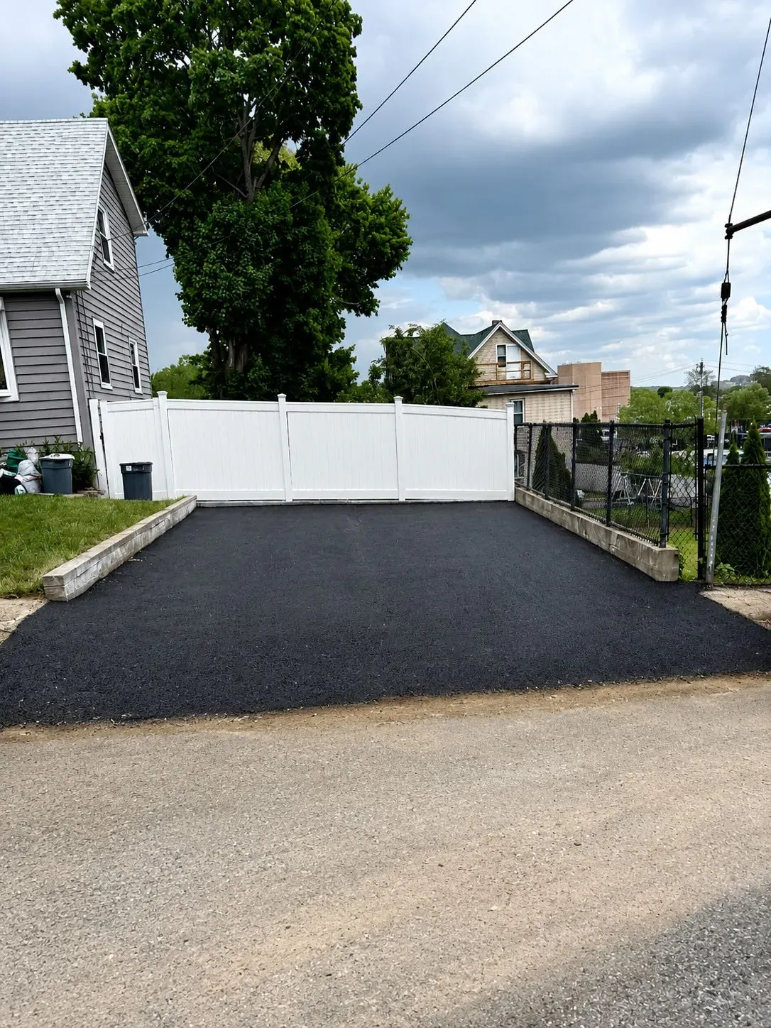 Fresh black asphalt driveway leading to a white gate, bordered by a chain-link fence and house siding.