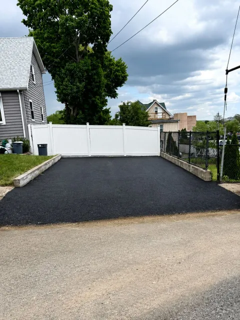 Fresh black asphalt driveway leading to a white gate, bordered by a chain-link fence and house siding.