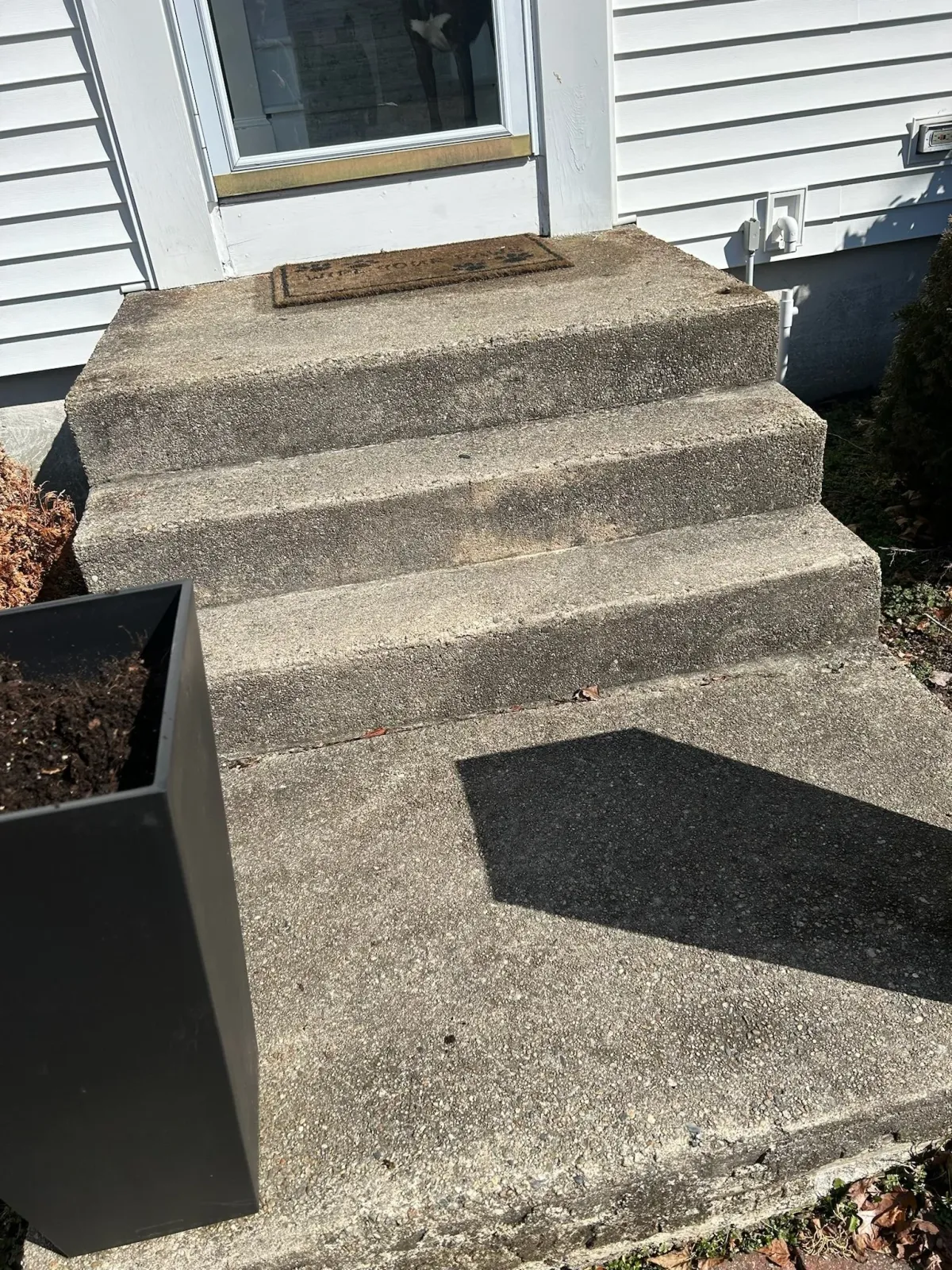 Concrete steps leading up to a white house door, with a doormat and a black planter box on the left.