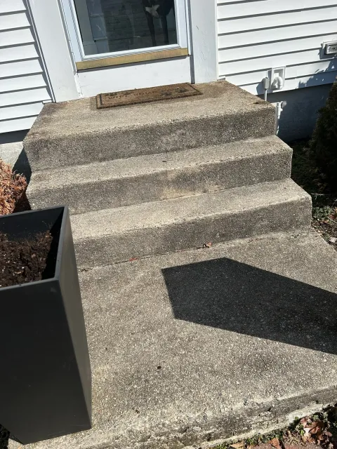 Concrete steps leading up to a white house door, with a doormat and a black planter box on the left.