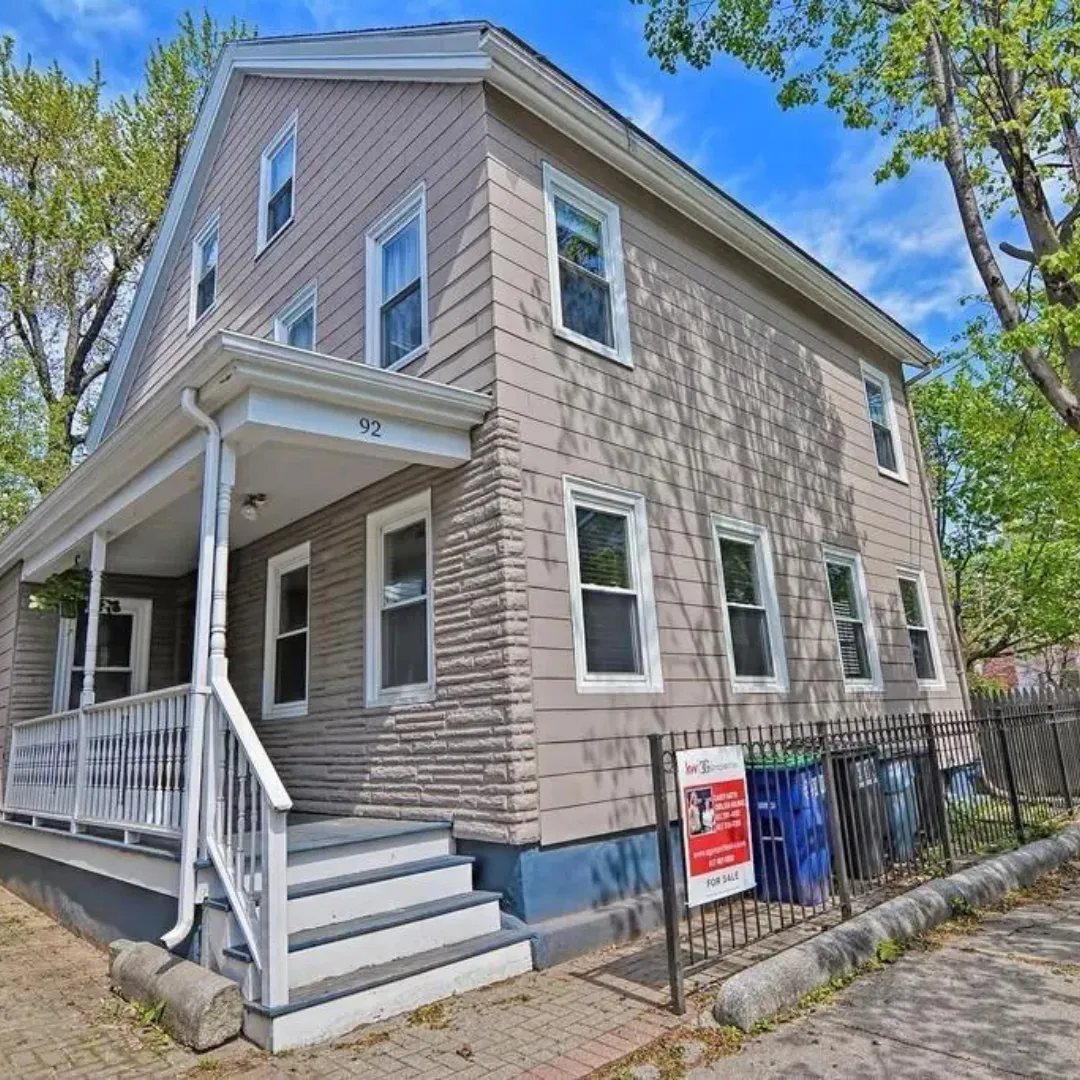 A three-story beige house with white trim, a small front porch with stairs, several windows, and a fenced yard with a red 'For Sale' sign.