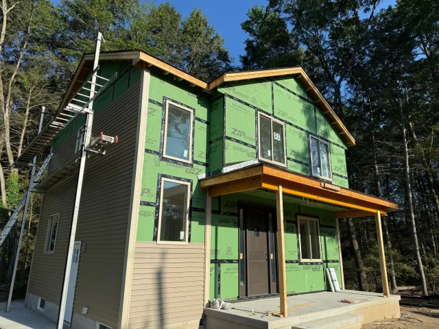 Two-story house under construction, green sheathing, brown siding, scaffolding, forest background.
