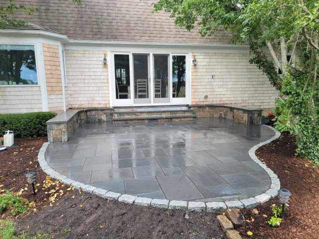 Wet outdoor patio area with dark gray stone tiles, a stone border, and steps leading to a house with sliding glass doors. Surrounding it are flower beds with mulch and small garden lights.