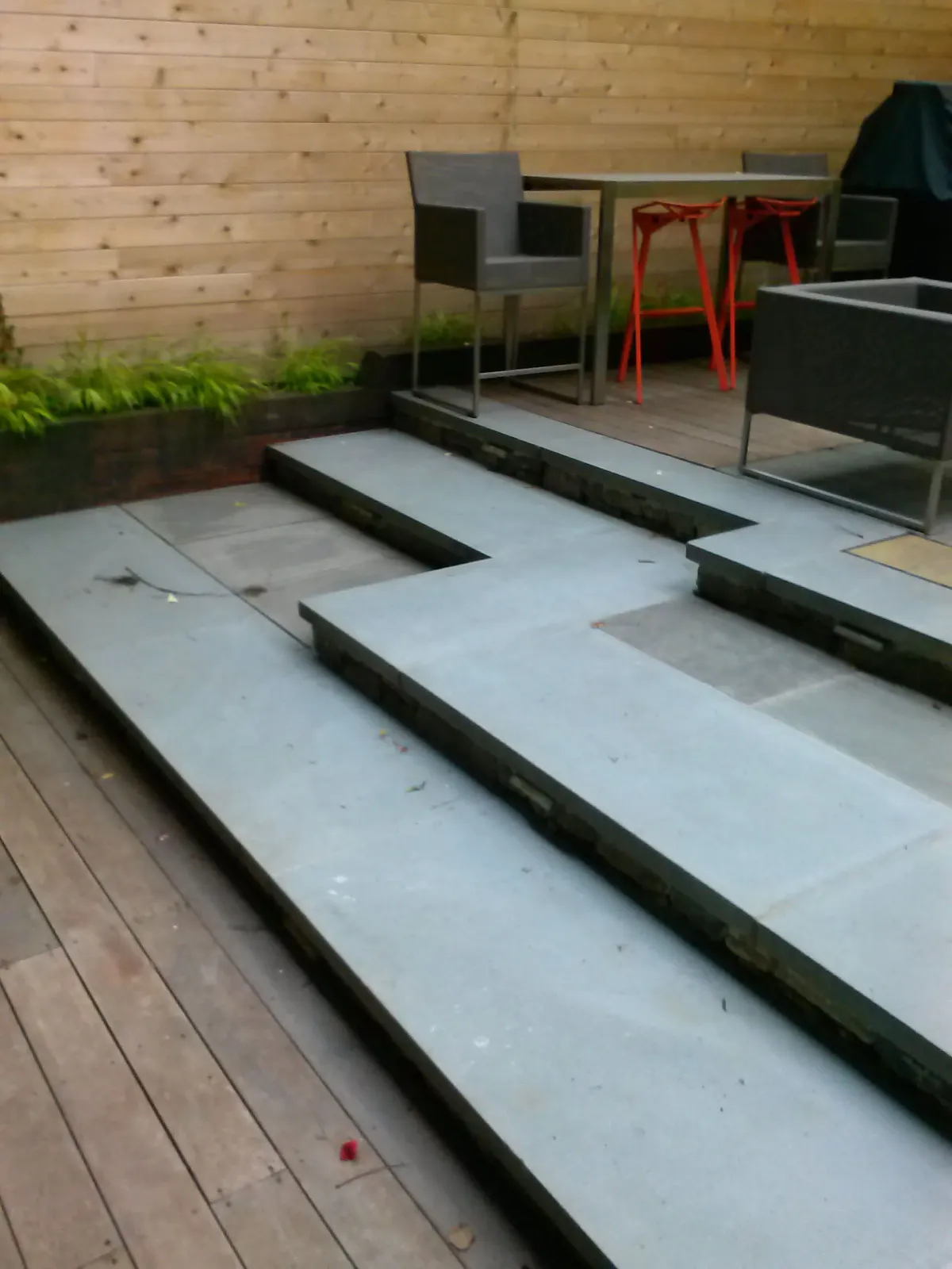 Outdoor patio with stepped concrete seating, wooden deck, and metal chairs against a wood wall