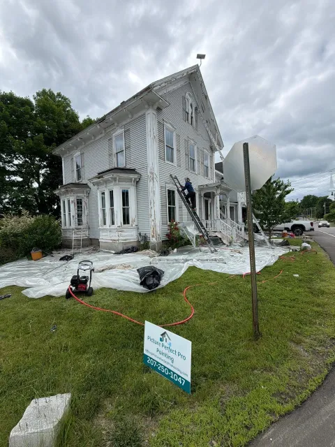 Damaged white house with debris and equipment in yard, likely after storm or renovation.