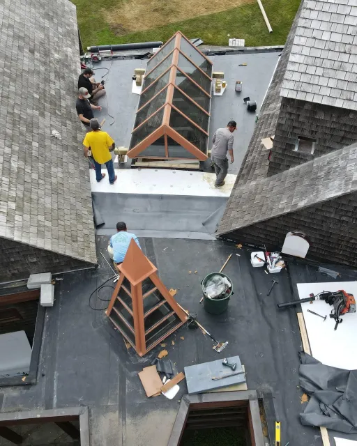 Roof workers assembling wooden skylight frames on a flat rooftop between slate buildings.