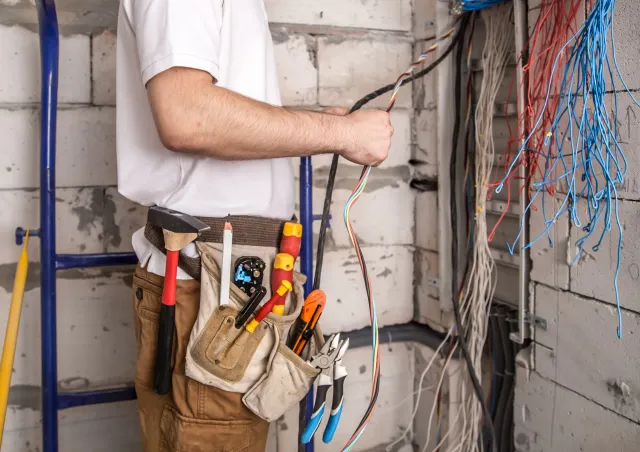 Electrician working on exposed wiring in a wall, with tools in a tan belt and blue cable bundles nearby