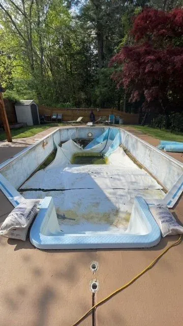 An empty, dirty swimming pool with a blue liner, surrounded by a concrete deck in a backyard with trees and a wooden fence.