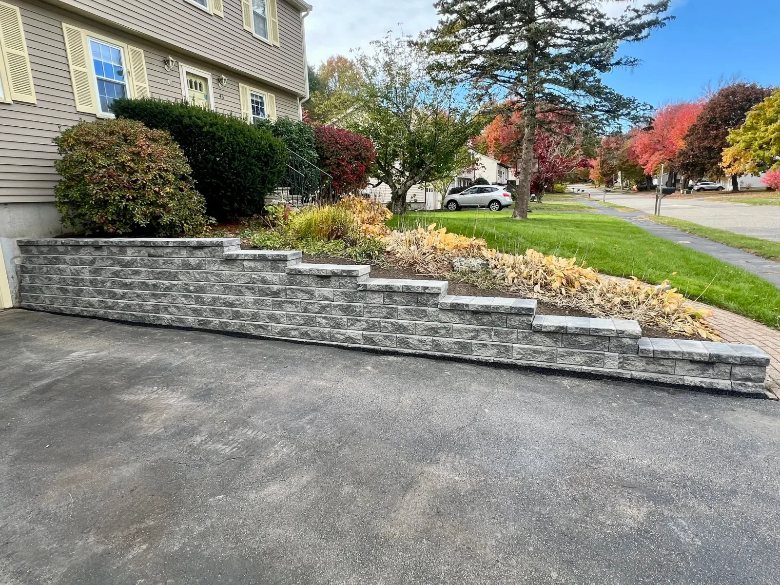 Retaining wall made of gray bricks next to a lawn with trees displaying fall colors, in a suburban neighborhood.