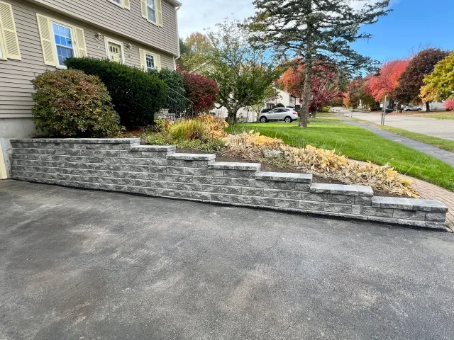 Retaining wall made of gray bricks next to a lawn with trees displaying fall colors, in a suburban neighborhood.