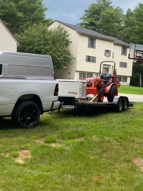 White pickup towing a trailer with an orange mower in a grassy yard beside a house