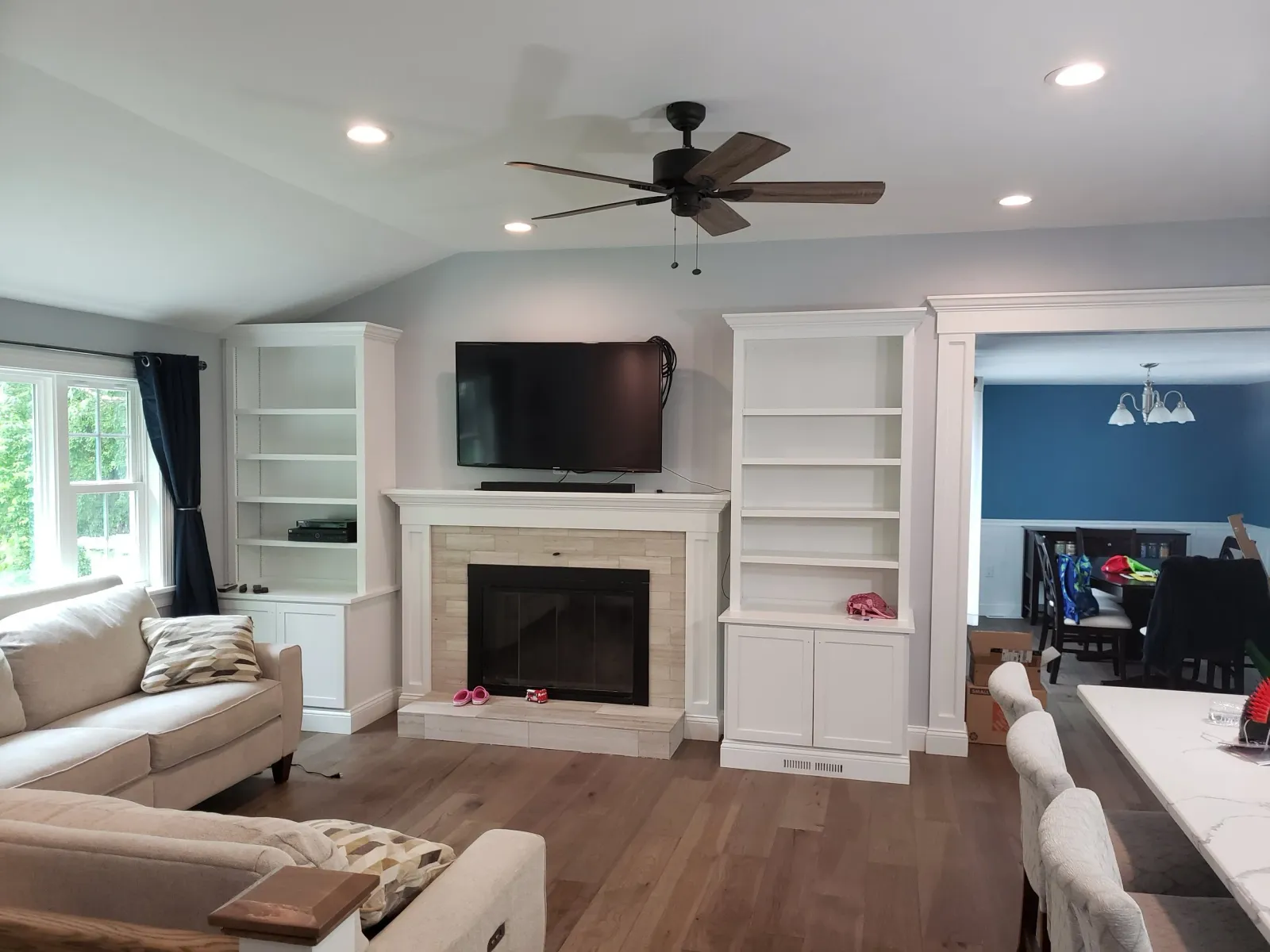 Living room with beige sofas, a fireplace with a mounted TV above, white built-in shelves, and a ceiling fan. Dining room with blue walls visible through a doorway.