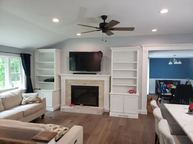 Living room with beige sofas, a fireplace with a mounted TV above, white built-in shelves, and a ceiling fan. Dining room with blue walls visible through a doorway.