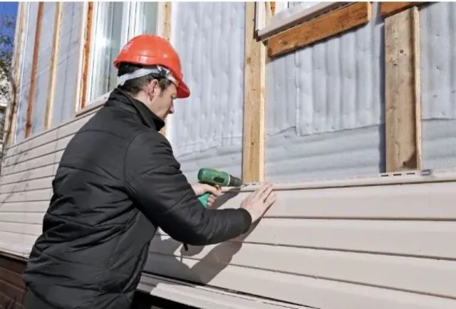 Construction worker installing siding with a power drill on a building exterior.