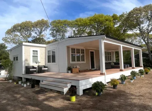 White tiny house with a large wooden deck and covered porch in a wooded yard