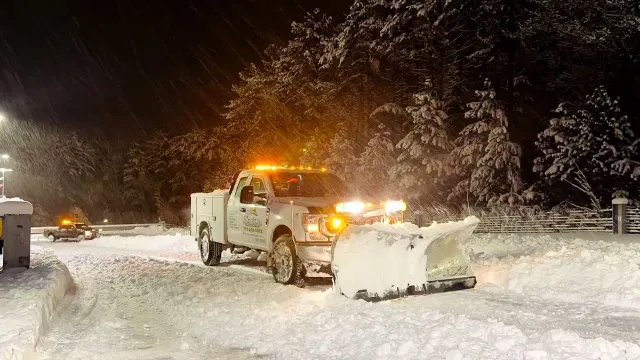 Snowplow clearing a snow-covered road at night, with bright lights and falling snow.