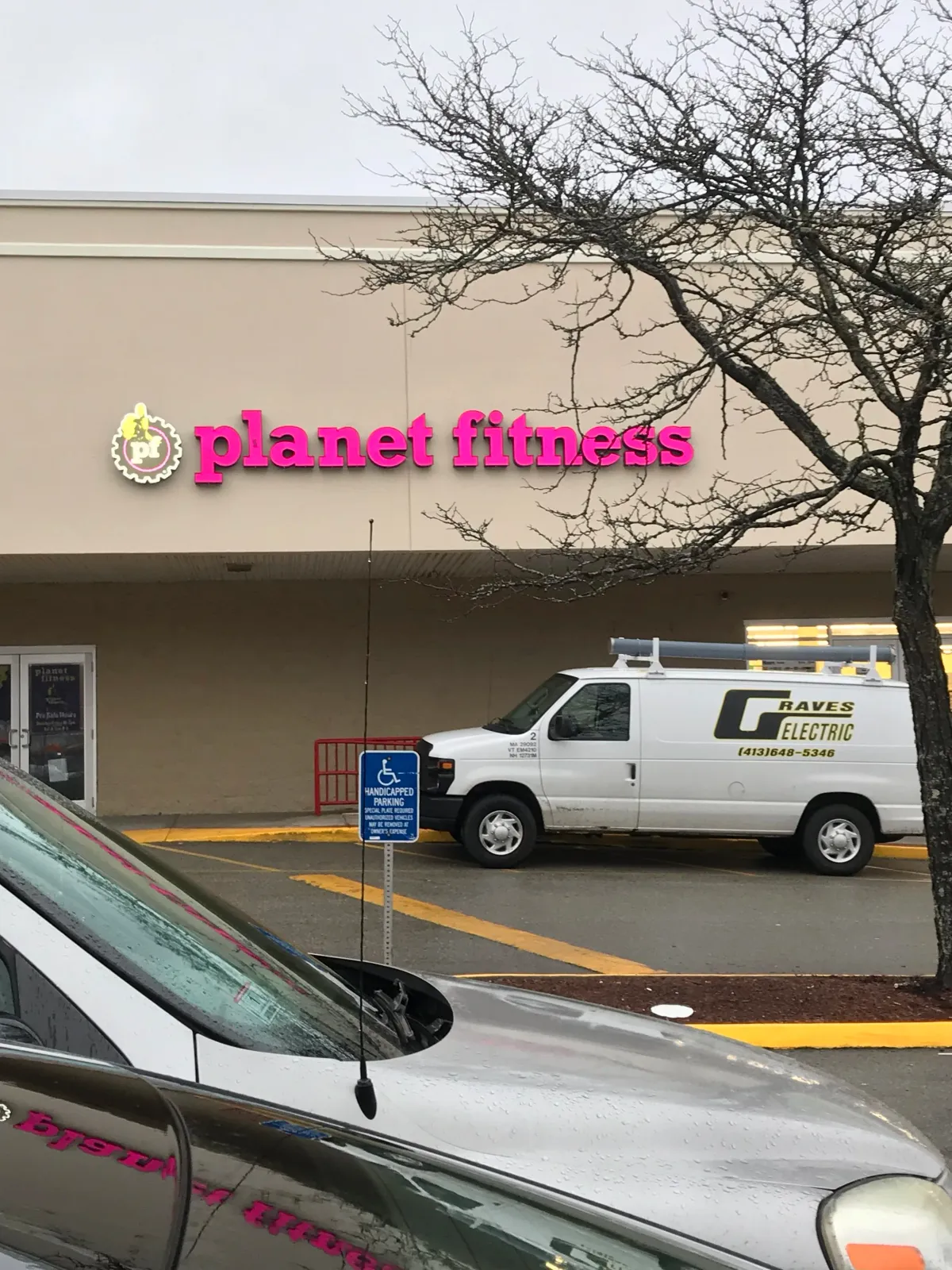 The image shows a Planet Fitness gym sign in pink letters on a beige building, with a tree in the foreground, a blue wheelchair accessible parking sign, and a white electric work van parked nearby.