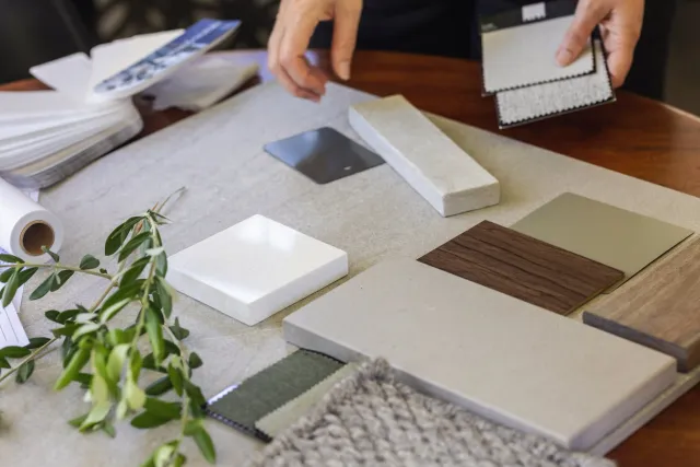 A person is holding two fabric samples, standing at a table with various material samples, including tiles, stones, and wood textures, with a plant in the foreground.