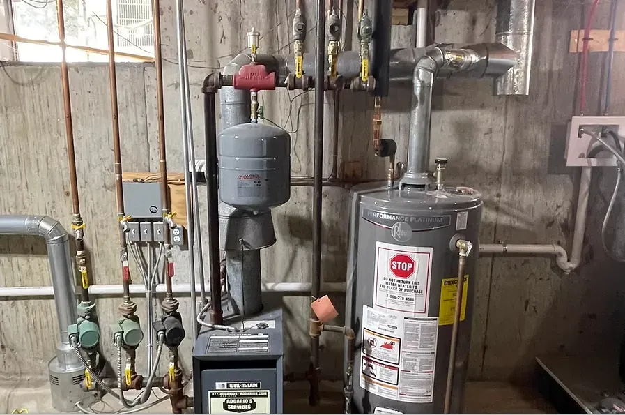 Basement utility area with water heater, pump, pipes, and wall-mounted electrical outlet.
