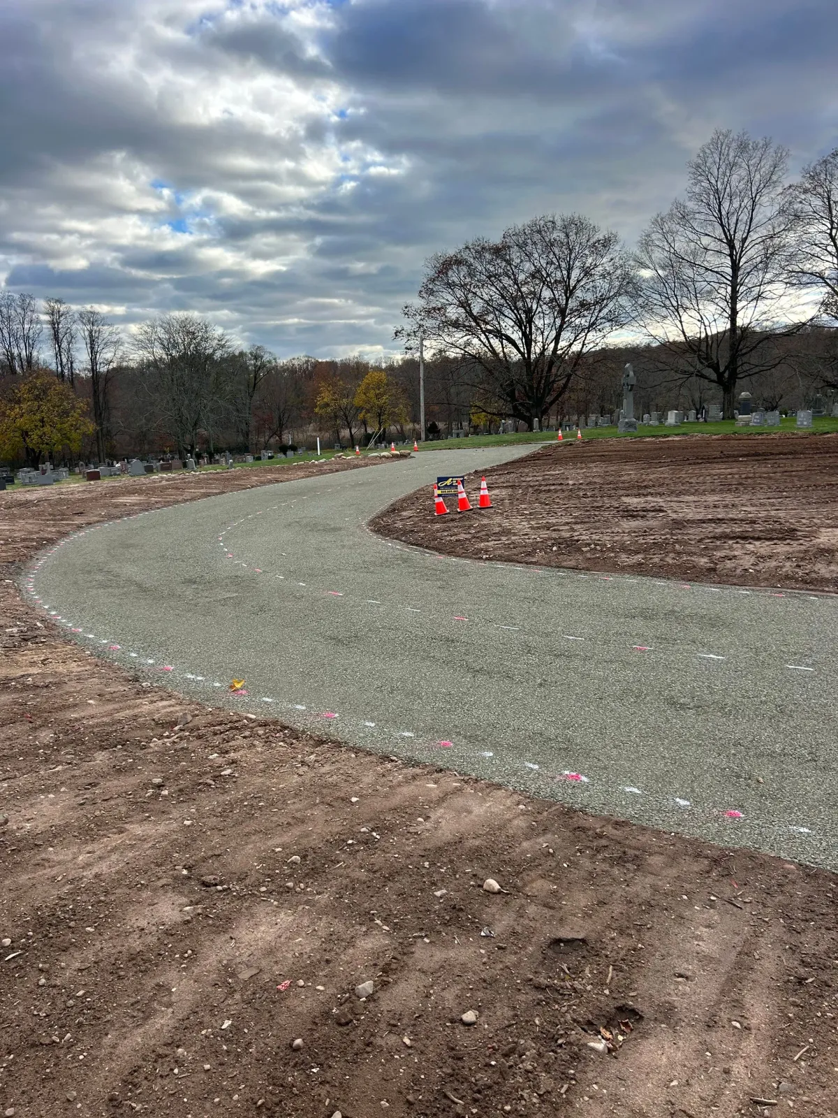 A curved gravel path winds through a cleared dirt construction site under a cloudy sky, with trees in the background.