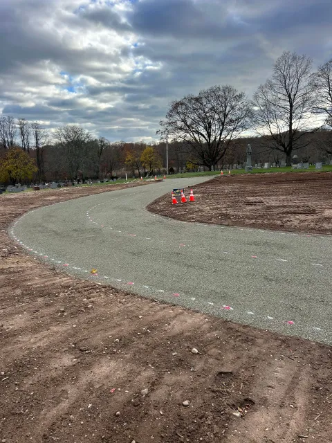 A curved gravel path winds through a cleared dirt construction site under a cloudy sky, with trees in the background.