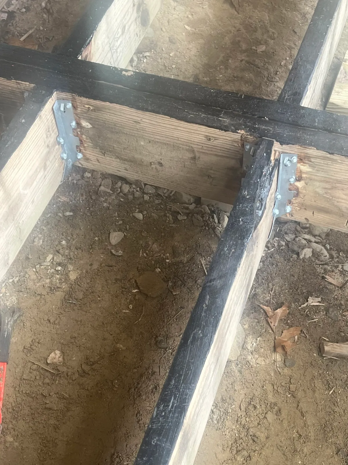 Close-up of a wooden and metal structure under construction, showing a wooden beam with metal brackets fastened with screws and bolts, with soil and dirt underneath.