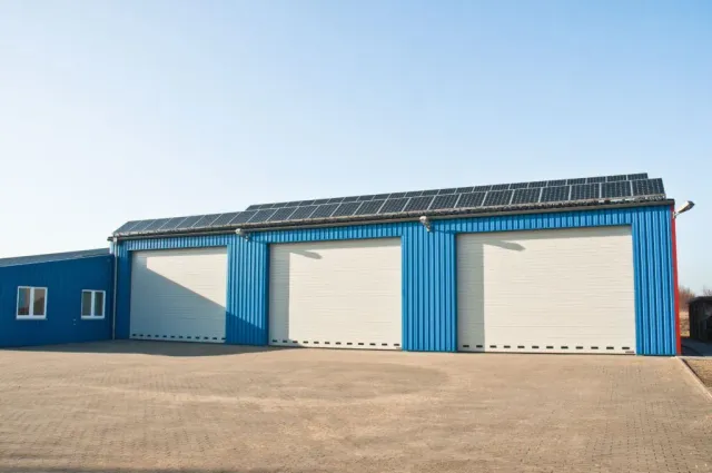 Blue industrial building with three garage doors, solar panels on the roof, sunny outdoor setting.