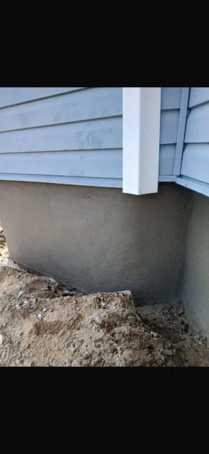 Exterior house corner with blue siding and stucco foundation. Dirt surrounds the foundation. White downspout.
