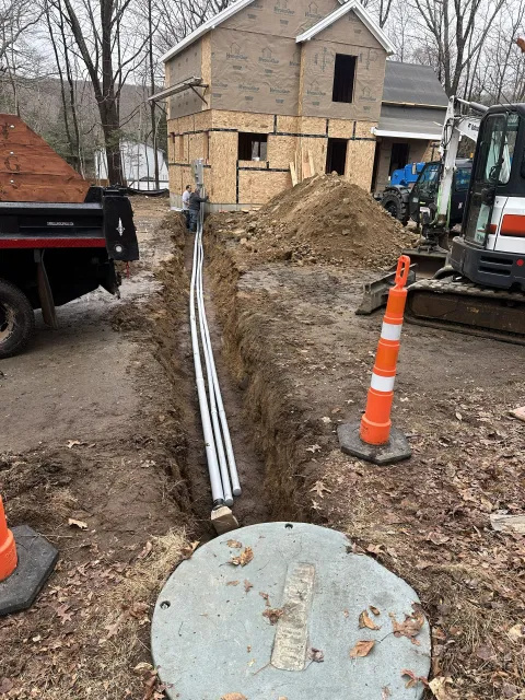 Trench with pipes, likely utilities, being installed near a house under construction. Excavator, truck, and traffic cones visible.
