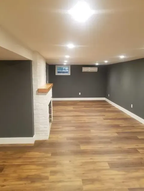 Newly renovated basement with wood-look flooring, gray walls, a white brick fireplace, and recessed lighting.