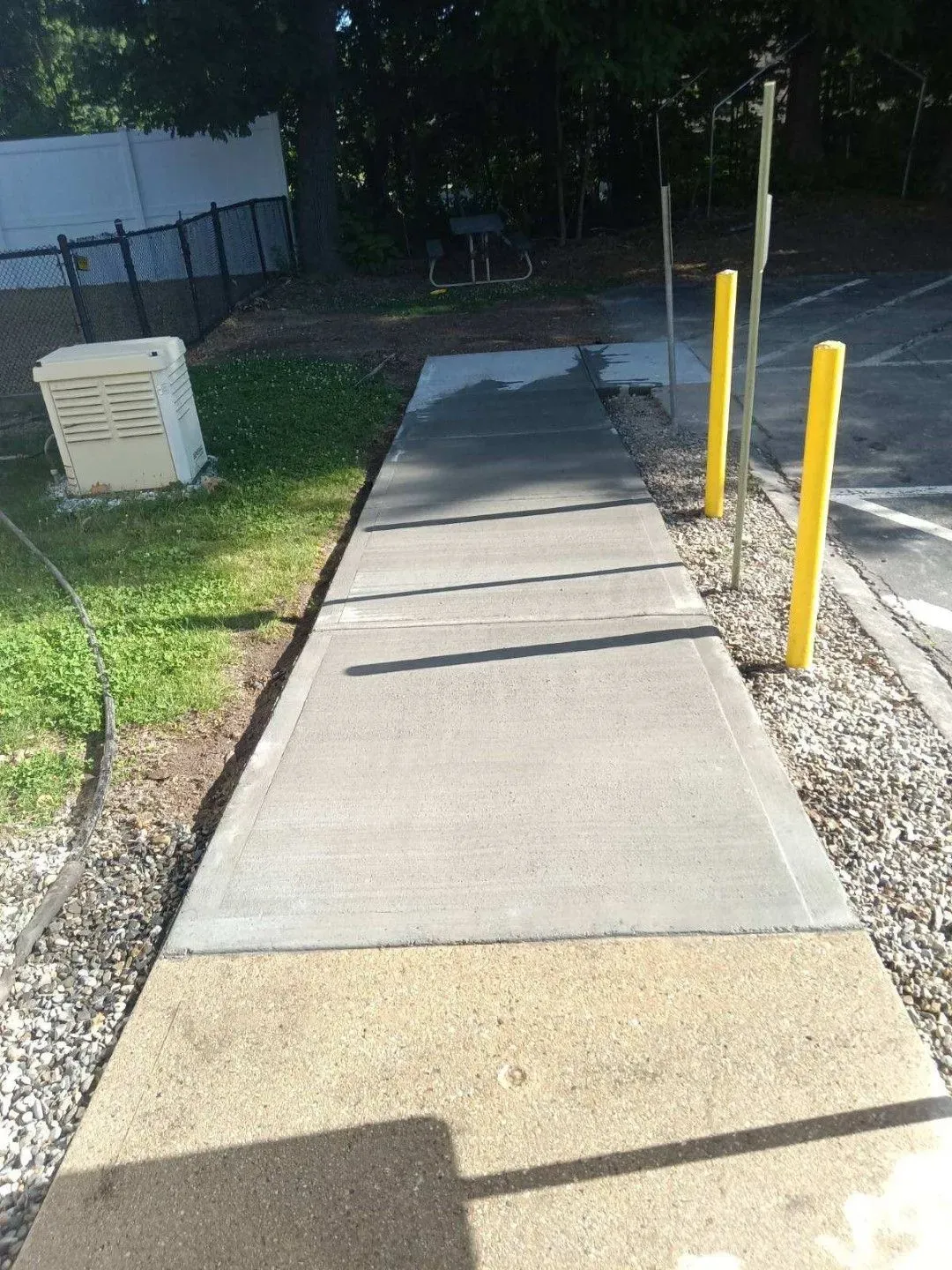 Concrete sidewalk with yellow bollards beside a grassy area and building wall, lit by sunlight and shadow.