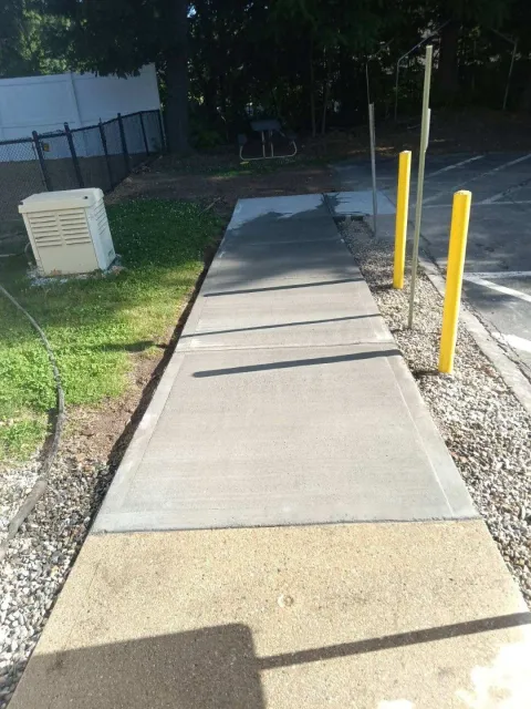 Concrete sidewalk with yellow bollards beside a grassy area and building wall, lit by sunlight and shadow.
