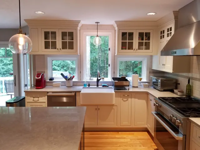 Modern kitchen with white cabinets, large windows overlooking greenery, stainless steel appliances, a white farmhouse sink, and pendant lighting.