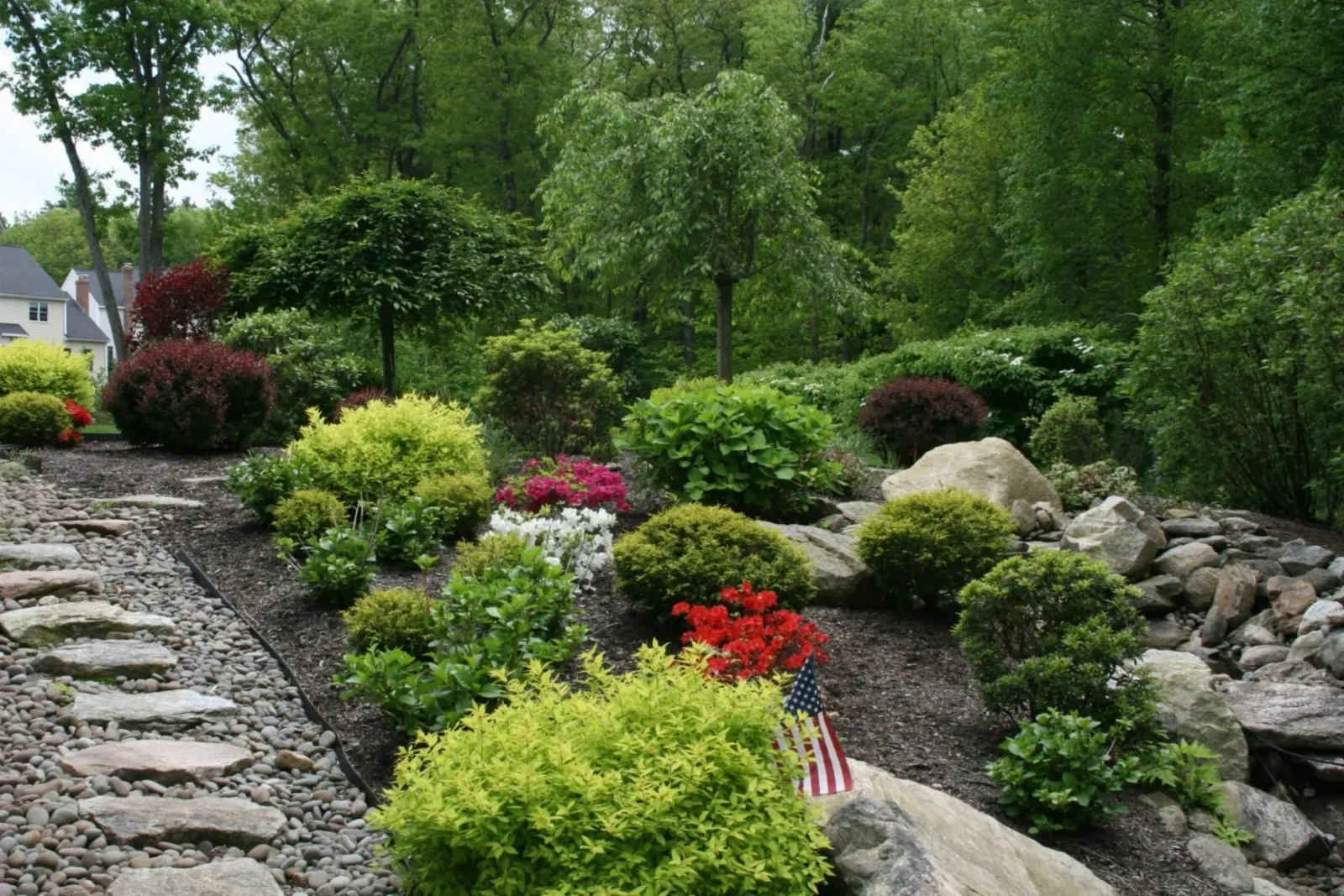 A landscaped garden with various shrubs, flowers, and a small tree. A stone path runs through the garden, and there is an American flag placed among the plants. Trees and greenery surround the area, and a house is partially visible in the background.