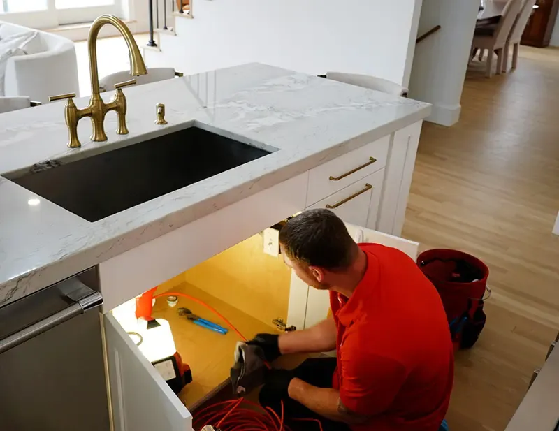 Plumber in red shirt working under kitchen sink, showcasing plumbing services for HVAC and plumbing needs in Taunton, MA.
