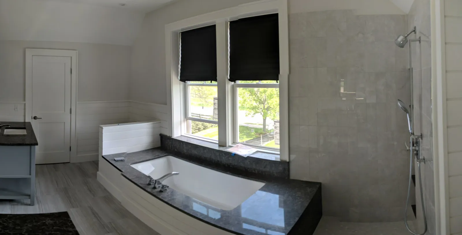 A modern bathroom with a built-in bathtub beneath two large windows with black shades, a walk-in shower with multiple showerheads, a granite countertop, and light-colored wooden paneling on the walls.