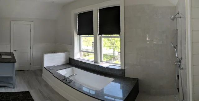 A modern bathroom with a built-in bathtub beneath two large windows with black shades, a walk-in shower with multiple showerheads, a granite countertop, and light-colored wooden paneling on the walls.